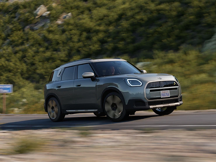מבט צדדי על ה-MINI Countryman החשמלית בנסיעה בצבע ירוק Smokey Green וגג בצבע כסף Vibrant Silver בשטח.