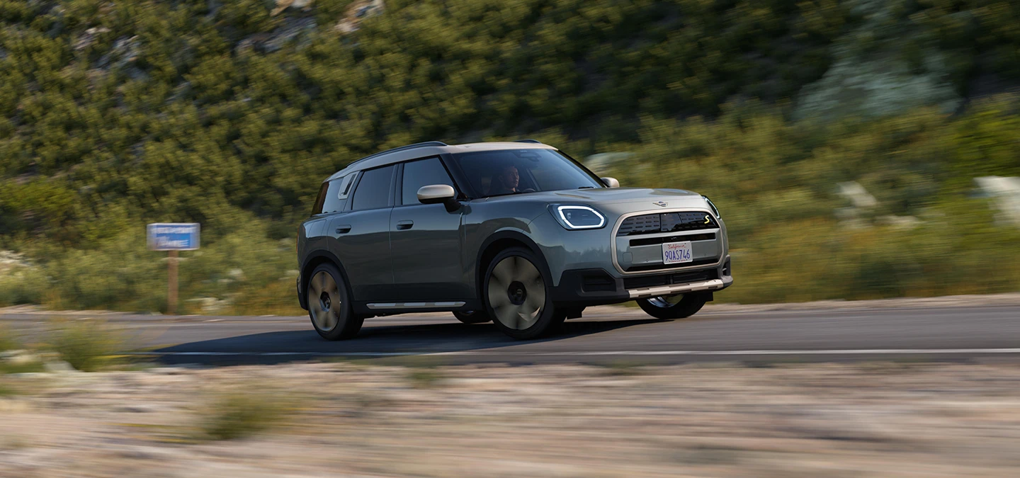 מבט צדדי על ה-MINI Countryman החשמלית בנסיעה בצבע ירוק Smokey Green וגג בצבע כסף Vibrant Silver בשטח.