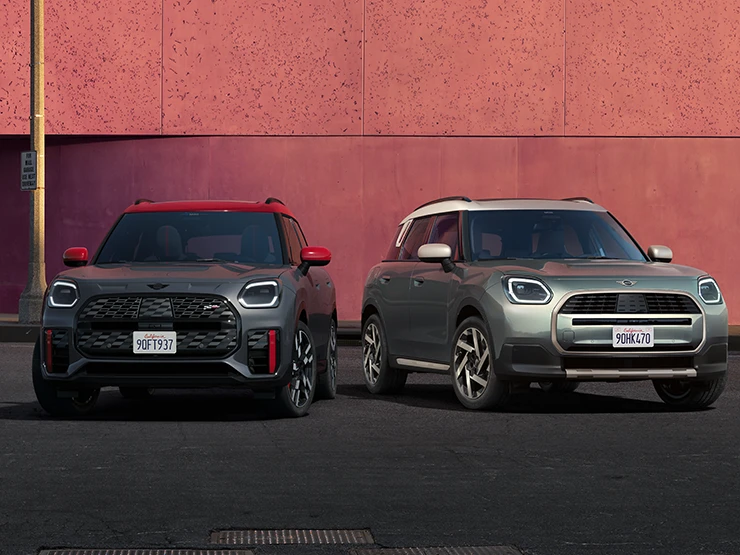 מבט קדמי של ה-MINI Countryman JCW בצבע אפור Legend Grey וגג אדום ו-MINI Countryman בצבע ירוק Smokey Green וגג בצבע כסף Vibrant Silver עומדים זה לצד זה.