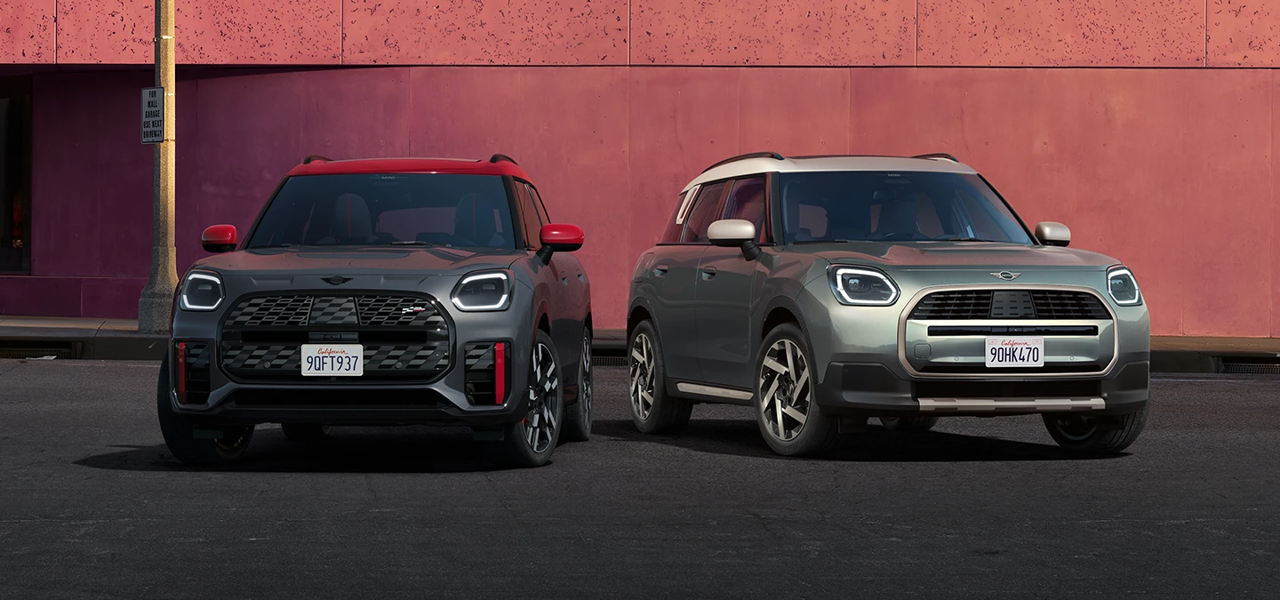 מבט קדמי של ה-MINI Countryman JCW בצבע אפור Legend Grey וגג אדום ו-MINI Countryman בצבע ירוק Smokey Green וגג בצבע כסף Vibrant Silver עומדים זה לצד זה.